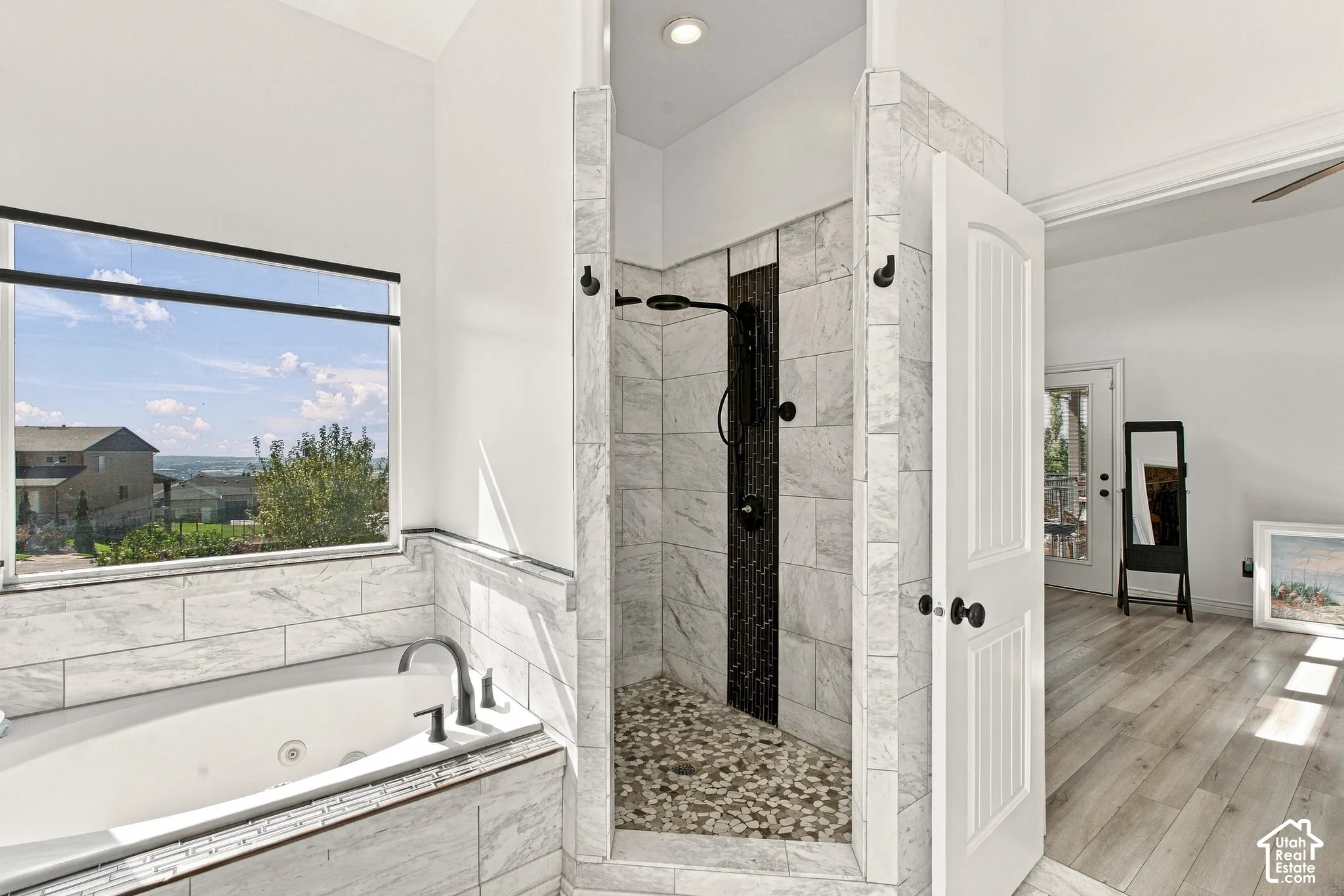 Jetted soaking tub with a tiled surround, walk-in shower featuring a river rock floor, black-finish shower fixtures, and wood-finish flooring