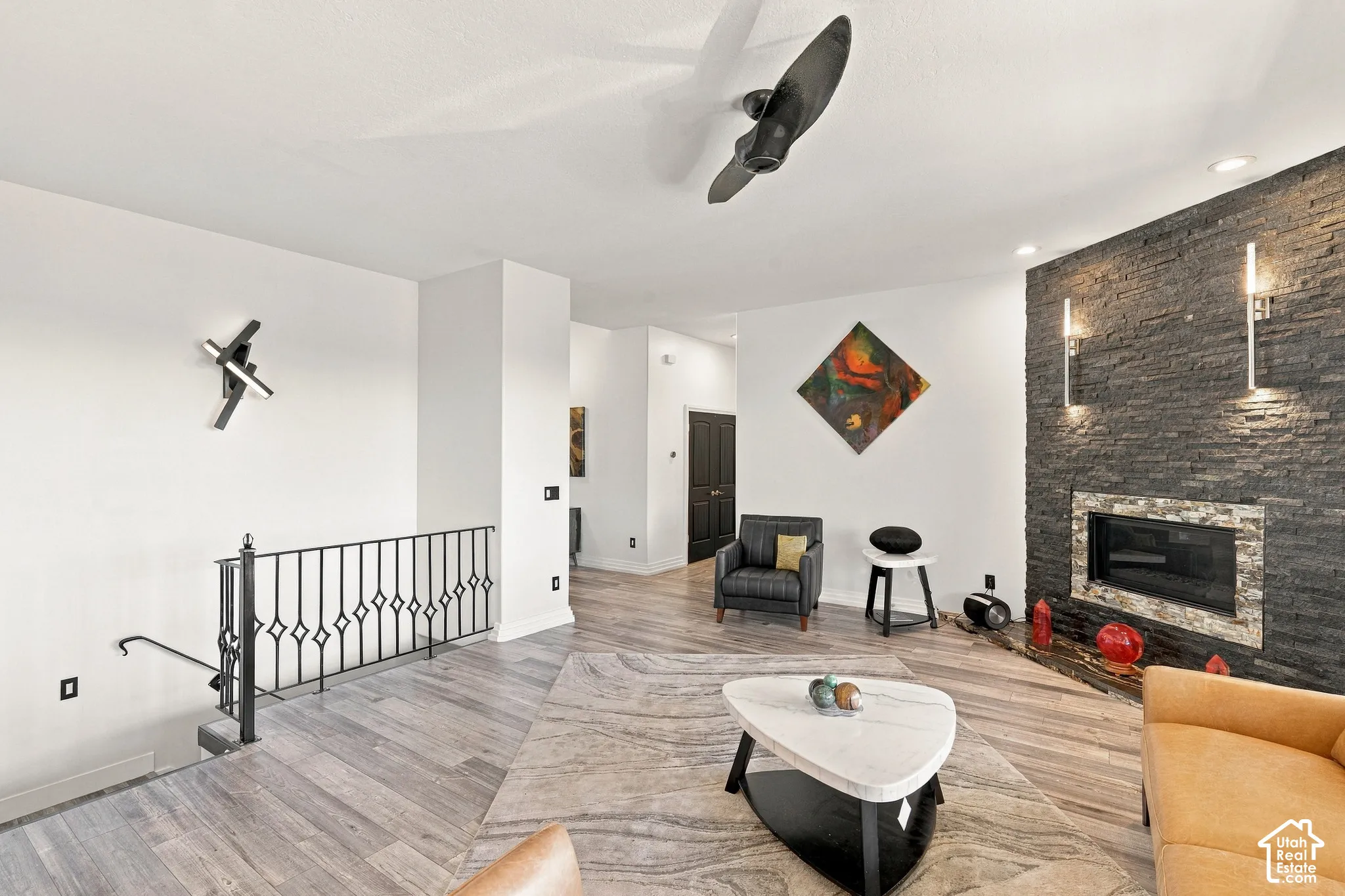 Wood-finish flooring, a stone-clad fireplace with sconce lighting, an open staircase with a decorative metal railing, and a ceiling fan