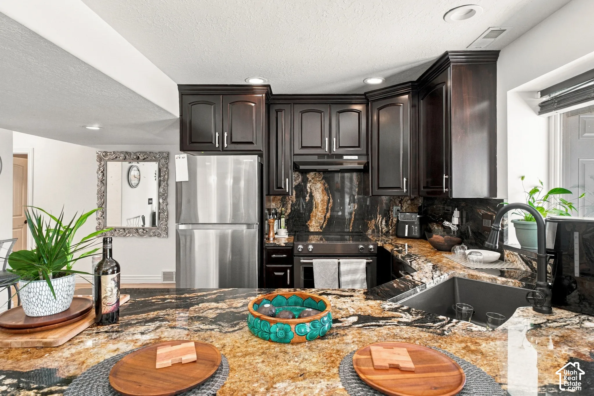 Kitchen featuring dark wood cabinetry, stainless steel appliances, a deep basin sink with a dark-finish faucet, and expansive natural stone countertops with a matching backsplash