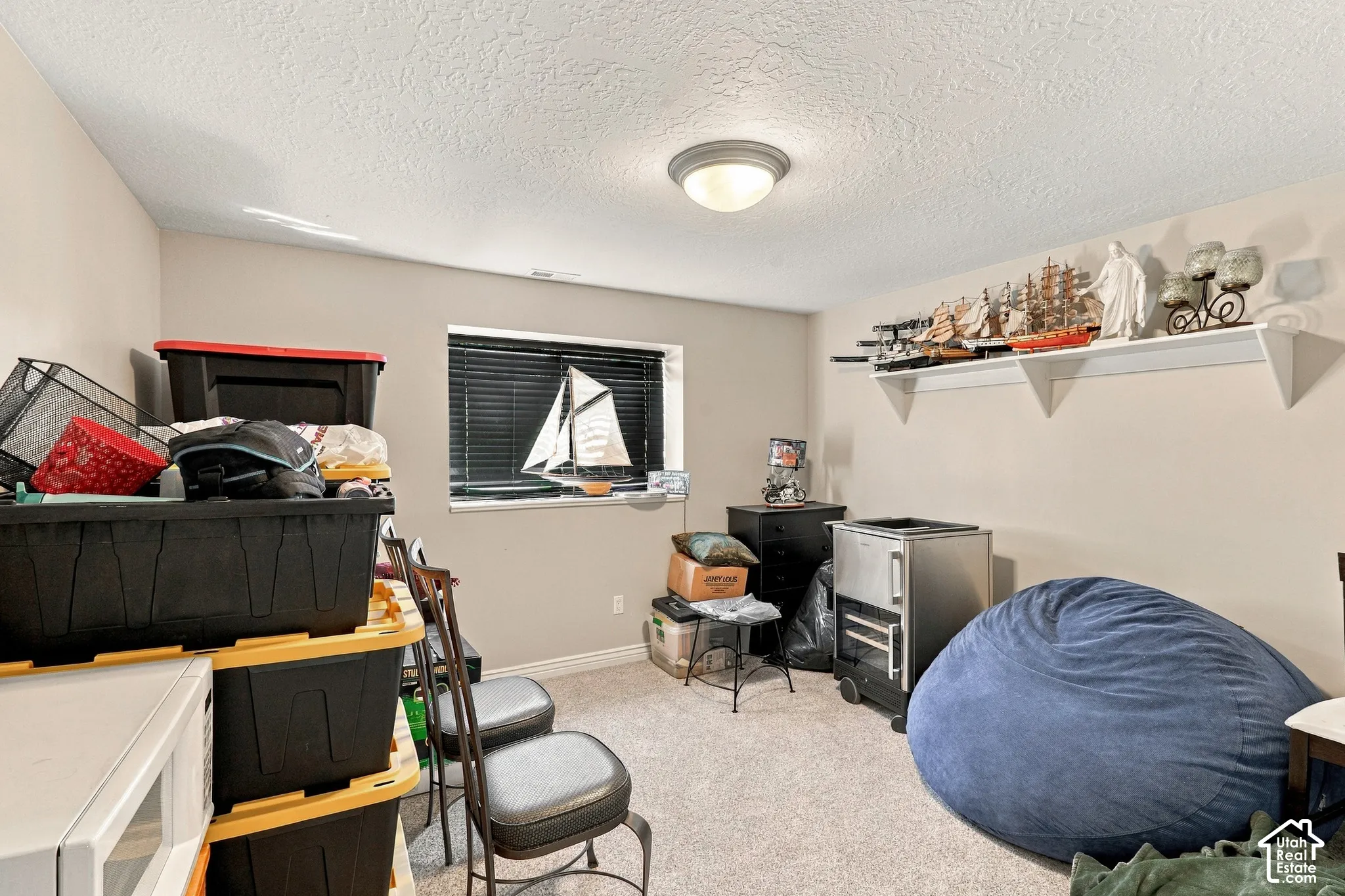 Carpeted flooring with a single window featuring horizontal blinds, a ceiling-mounted light fixture, and a wall-mounted display shelf