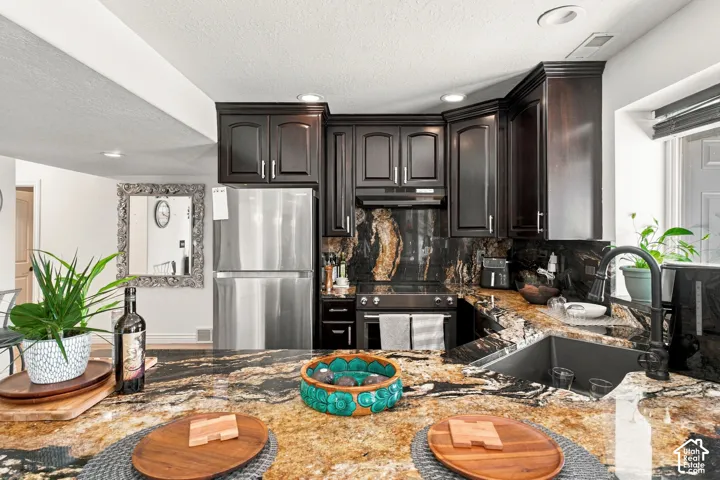 Kitchen featuring dark wood cabinetry, stainless steel appliances, a deep basin sink with a dark-finish faucet, and expansive natural stone countertops with a matching backsplash