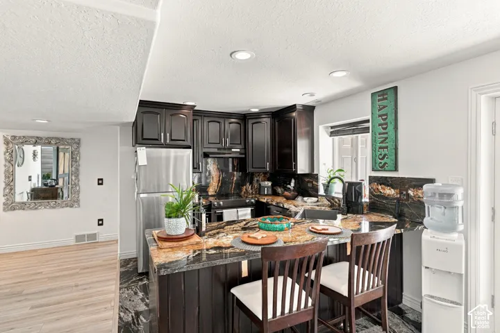 Kitchen featuring dark wood cabinetry, stainless steel appliances, and a multi-toned stone countertop with an integrated breakfast bar