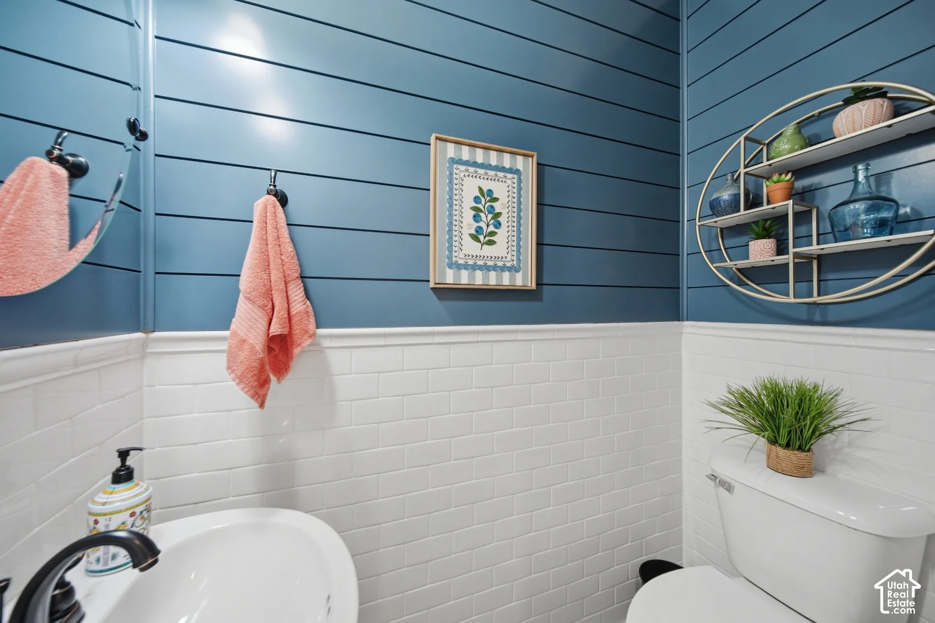 White subway tile wainscoting, blue shiplap walls, wall-mounted mirror, white pedestal sink with dark finish faucet, and white toilet
