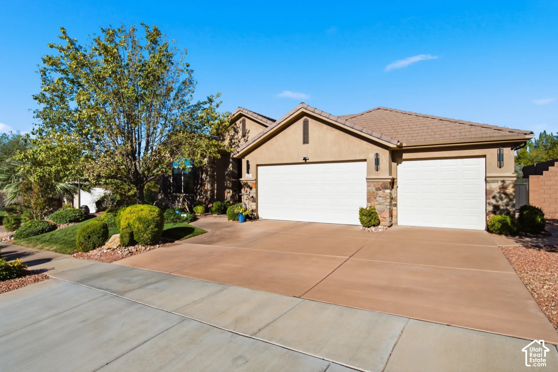 Stucco exterior with stone accents, a tile roof, two garage doors, a concrete driveway, and landscaping with mature greenery