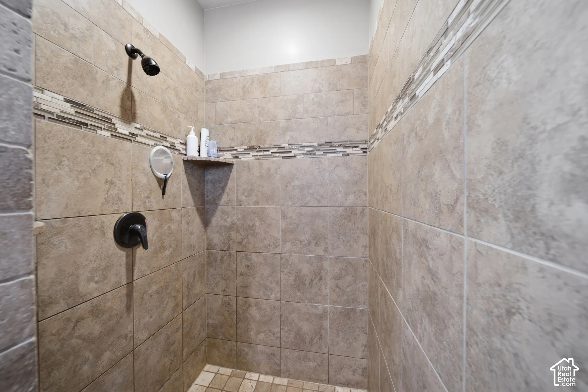 Tiled shower enclosure featuring a dark-finish shower head and control, a decorative mosaic tile accent band, and a built-in corner shelf