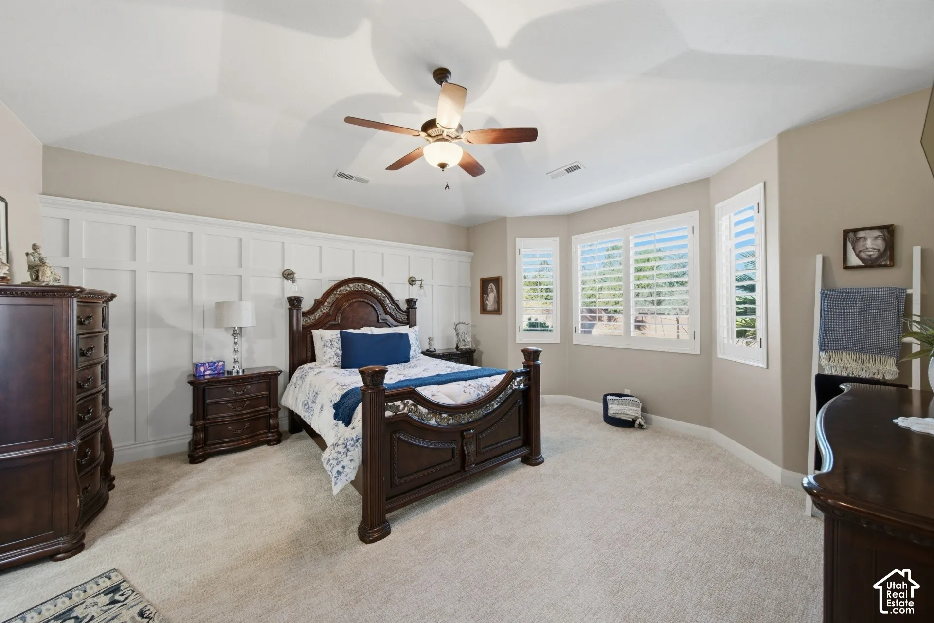 Tray ceiling with a ceiling fan, accent wall with wainscoting, and multiple windows with plantation shutters