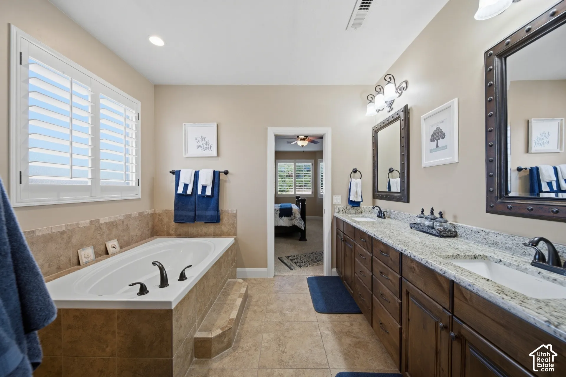 Built-in soaking tub with tile surround, white plantation shutters, dual sink vanity with stone countertop, and wood-finish cabinetry