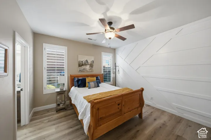 Accent wall with geometric paneling, two windows with plantation shutters, wood-finish flooring, and a ceiling fan with integrated lighting
