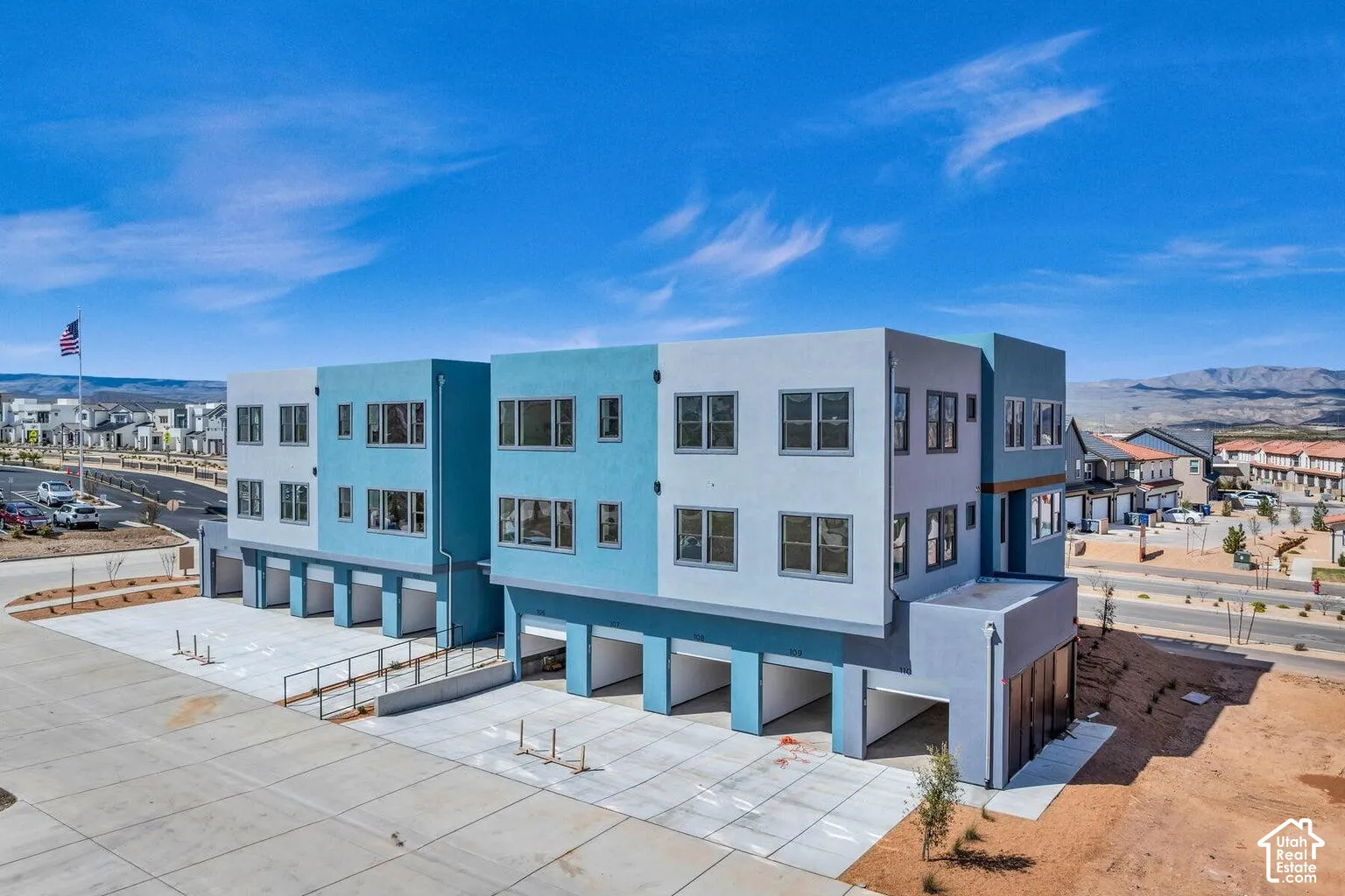Modern multi-story facade featuring distinct blue and grey color blocking, multiple large windows, ground-level covered parking, and a flat roofline