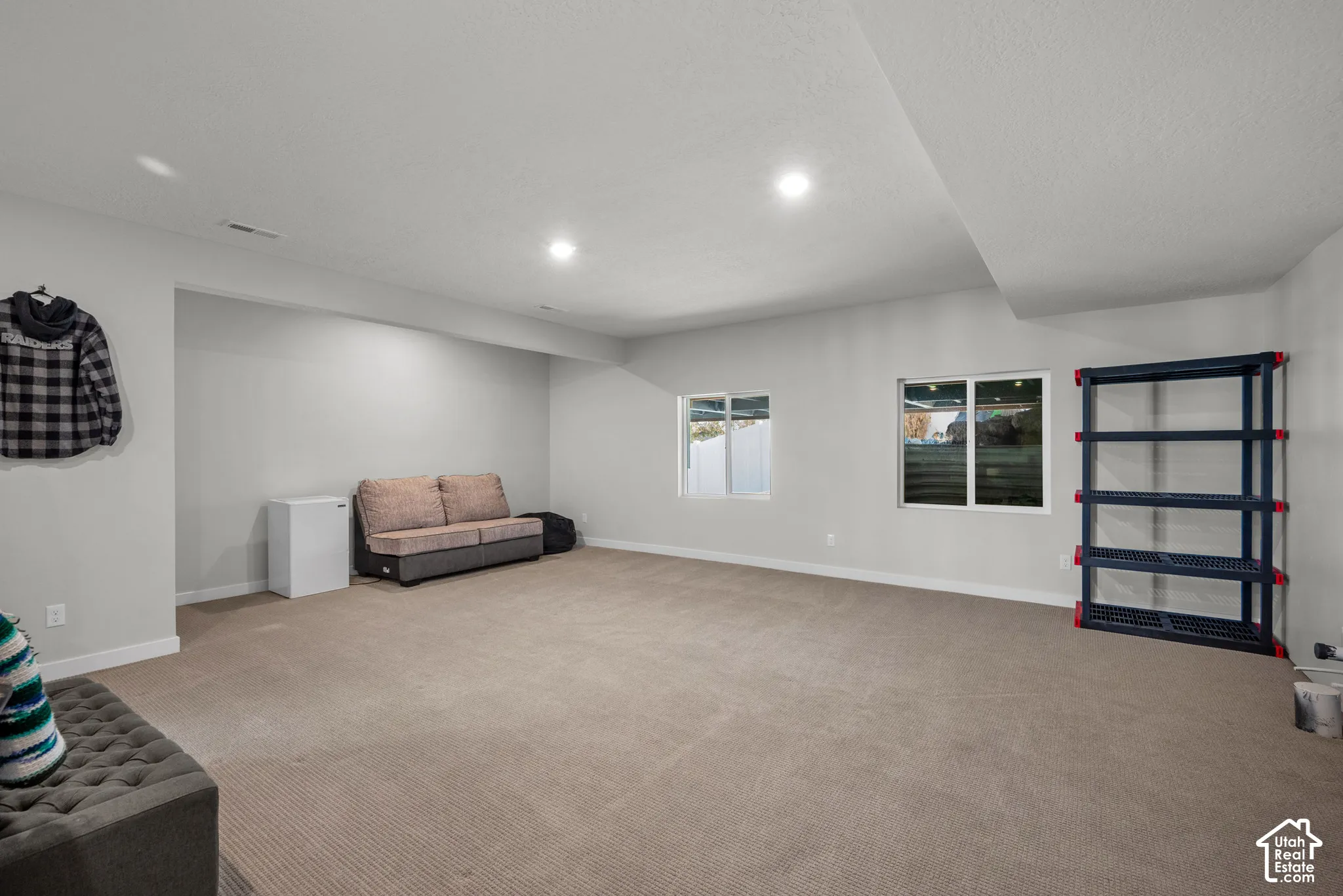 Recessed lighting illuminates a spacious room with light gray walls and neutral carpeting, featuring two windows