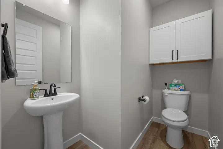 Pedestal sink with a matte black faucet, wall-mounted mirror, wood-finish flooring, white wall cabinets with dark hardware, and a white toilet