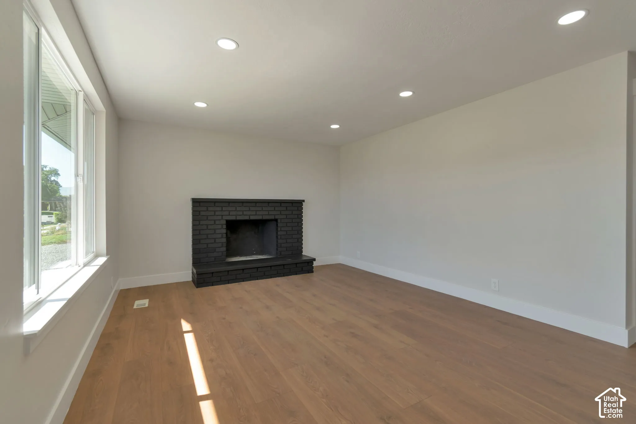 Spacious room with wood-finish flooring, a black brick fireplace, recessed lighting, and large windows