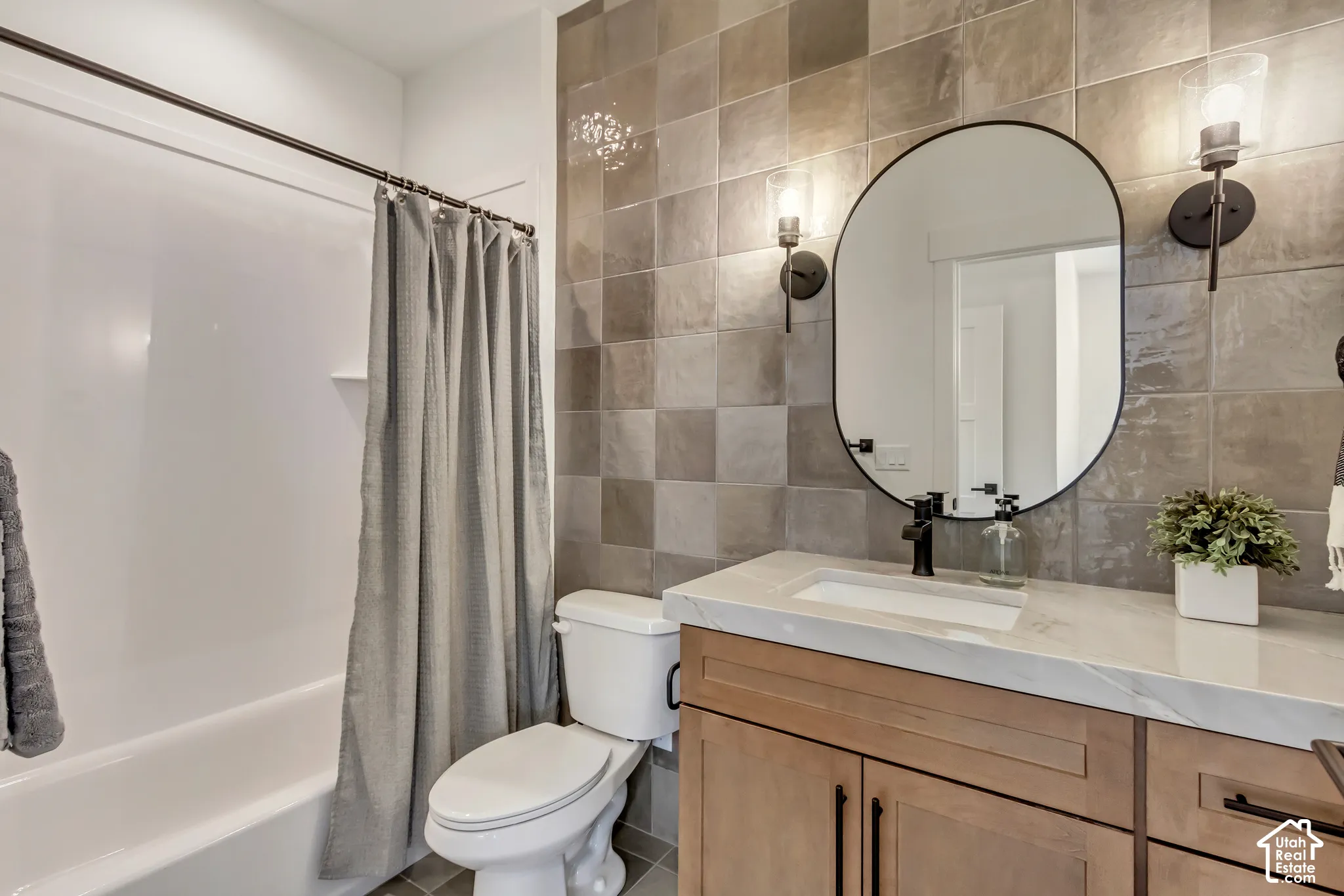 Built-in tub with shower, square wall tiles, light wood-finish vanity, white countertop with integrated sink, and black wall sconces