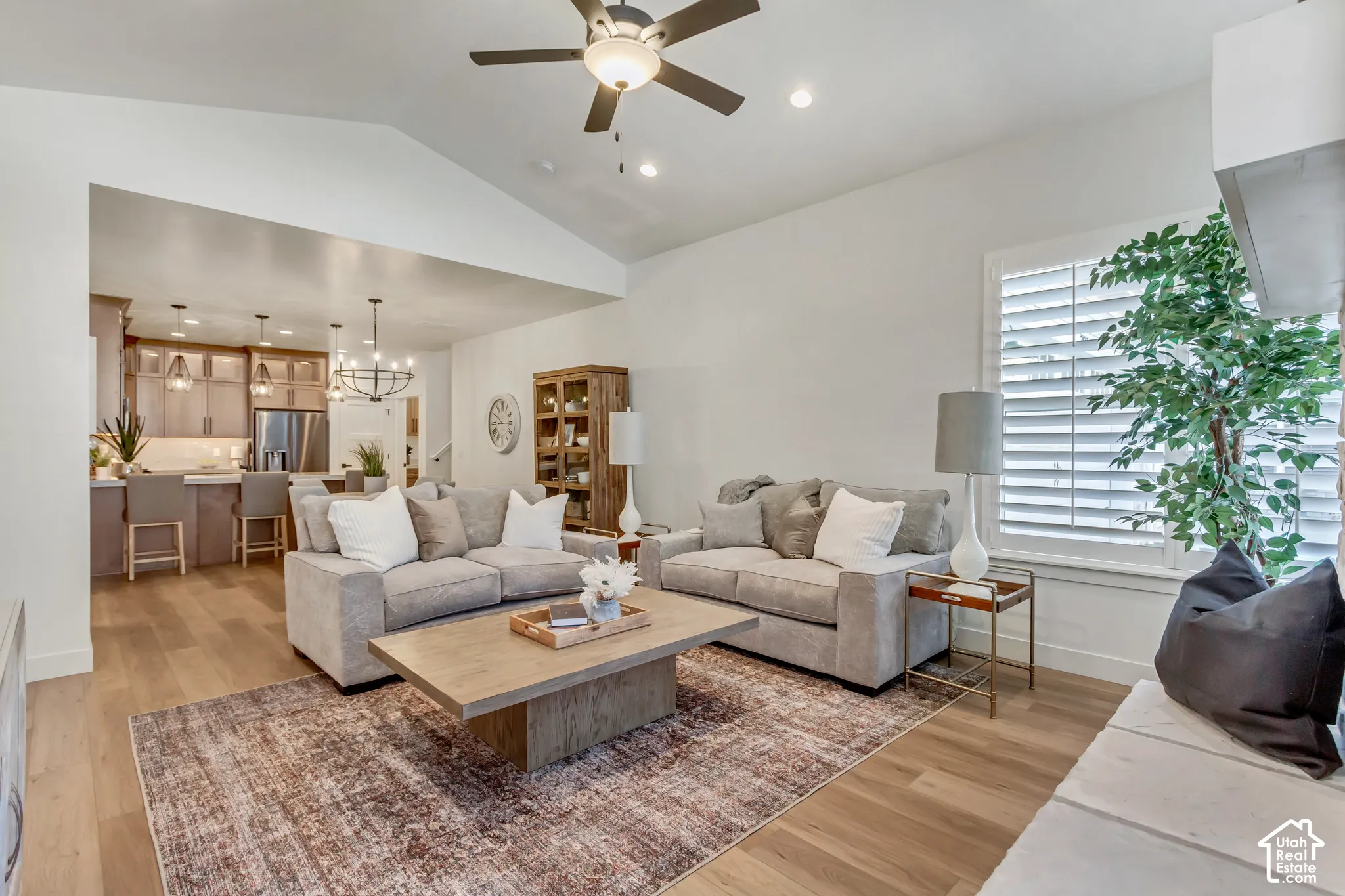 Vaulted ceiling with a ceiling fan, wide plank wood-finish flooring, white plantation shutters, and a built-in stone hearth