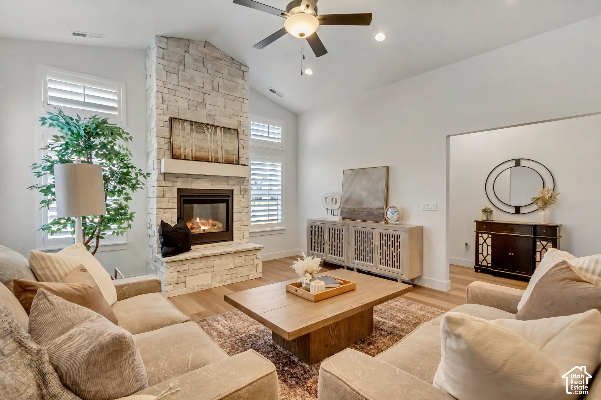Vaulted ceiling with a ceiling fan, stone-clad fireplace with a hearth and mantel, wood-finish flooring, and plantation shutters