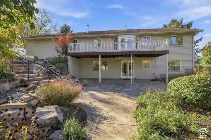 Rear exterior featuring a tiered stone staircase with metal railing, a covered concrete patio, an elevated wood-finish deck with railings, and established landscaping