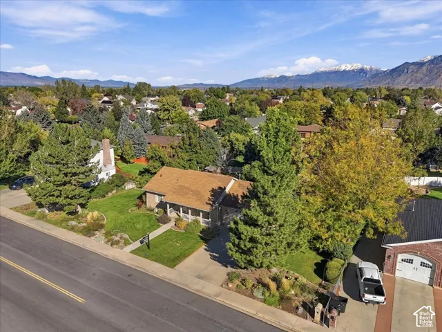 Single-story residence featuring a shingled roof, front-facing covered porch, concrete driveway, and expansive lawn with mature trees set against a mountain backdrop