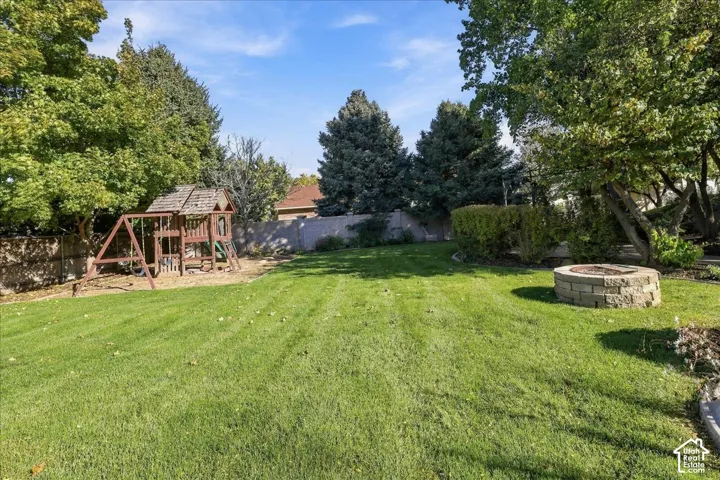 Backyard featuring a manicured lawn, stone fire pit, wood-frame play structure, mature trees, and perimeter fencing