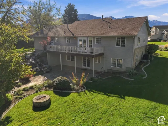 Rear exterior featuring a raised balcony with white railings, a ground-level stone patio, a circular stone fire pit, and a mountain backdrop