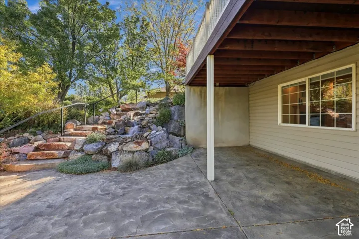 Walkout basement leads to covered deck, concrete patio featuring tiered stone landscaping, a built-in water feature, and a staircase with a metal handrail