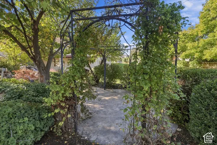 Ornate metal garden arbor featuring an arched frame and stone paver flooring, surrounded by mature trees, shrubs, and climbing greenery