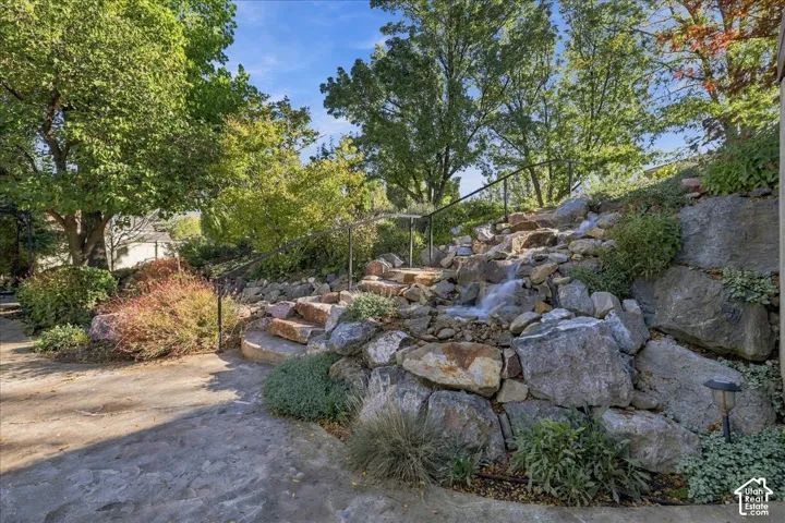 Landscaped backyard featuring a terraced stone retaining wall with a built-in water feature, flagstone hardscaping, metal handrails, and mature trees