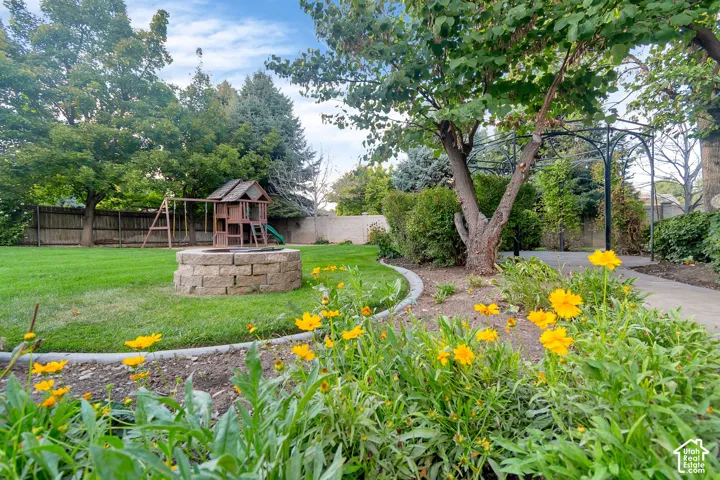 Expansive backyard featuring a stone fire pit, lush lawn, mature trees, and a decorative garden arbor