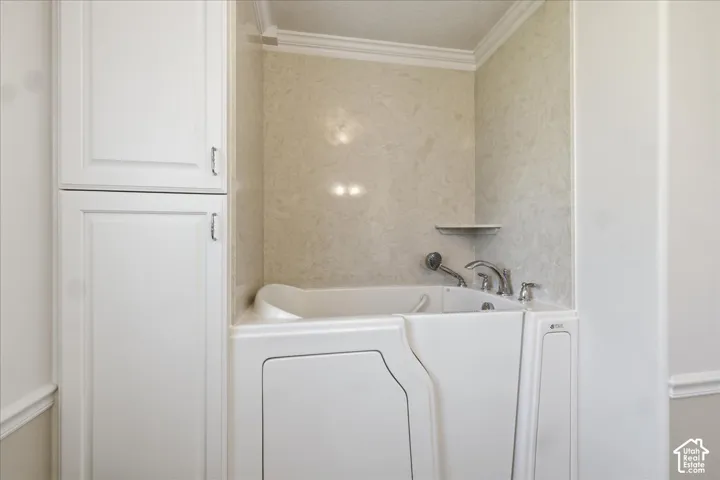 Primary bath. Walk-in tub featuring integrated chrome fixtures, a handheld showerhead, a built-in corner shelf, and surrounding wall panels, complemented by white floor-to-ceiling cabinetry with crown molding