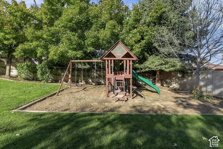 Rear yard featuring a manicured lawn, perimeter fencing, mature trees, and a wooden play structure with an integrated slide