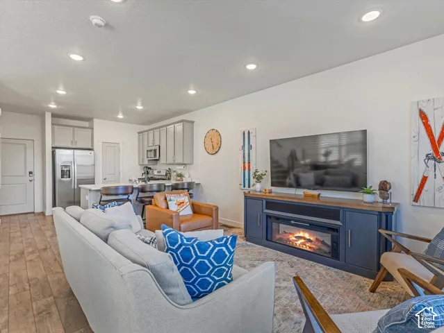 Open-concept living area featuring recessed lighting, wood-finish flooring, a kitchen island with seating, stainless steel appliances, and a built-in entertainment console with an electric fireplace