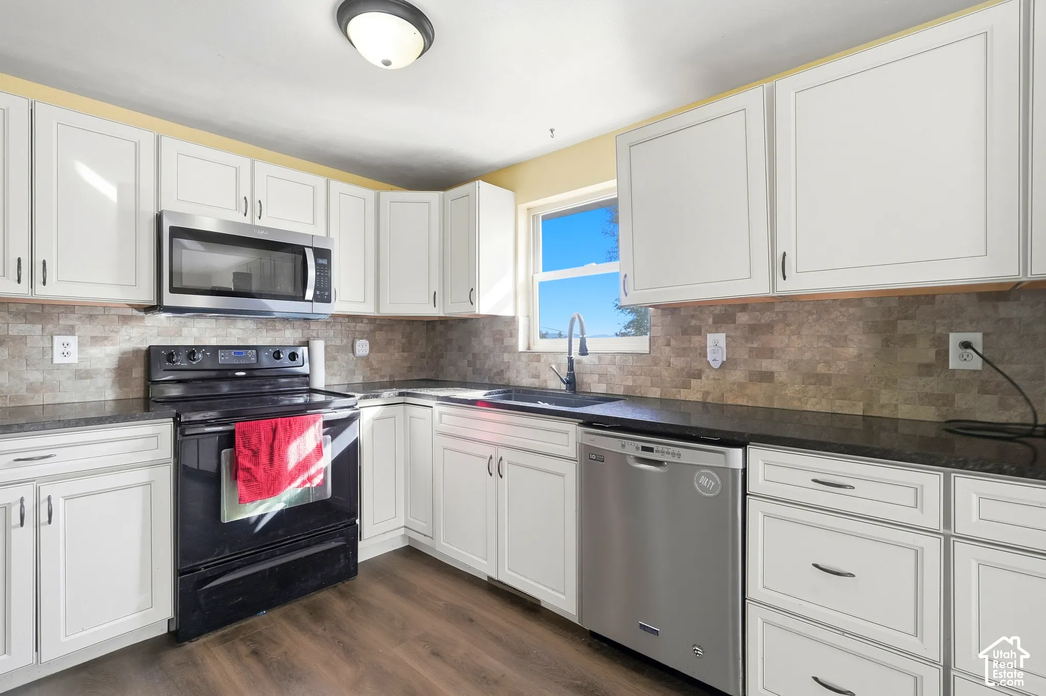 White cabinetry with dark hardware, dark countertops, a tiled backsplash, wood-finish flooring, and a stainless steel dishwasher
