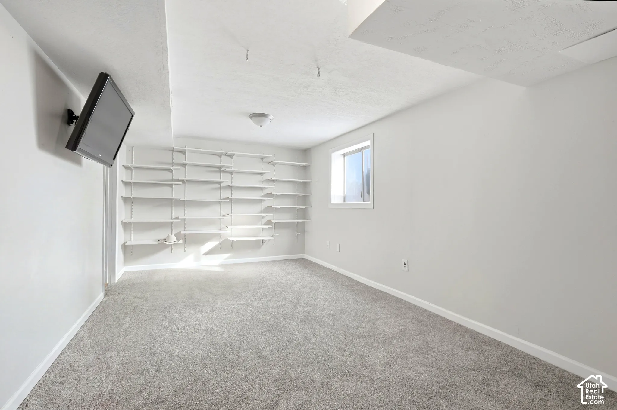 Wall-mounted shelving system, recessed window, wall-mounted television bracket, neutral carpet flooring, and flush-mount ceiling light fixture