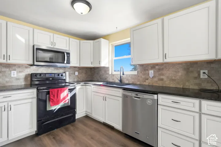 White cabinetry with dark hardware, dark countertops, a tiled backsplash, wood-finish flooring, and a stainless steel dishwasher