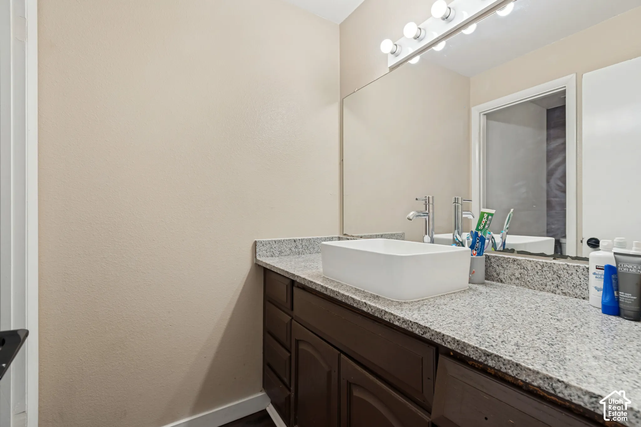 Vanity with a rectangular vessel sink, polished chrome faucet, granite-look countertop, dark wood-finish cabinetry, and a large wall-mounted mirror with integrated lighting