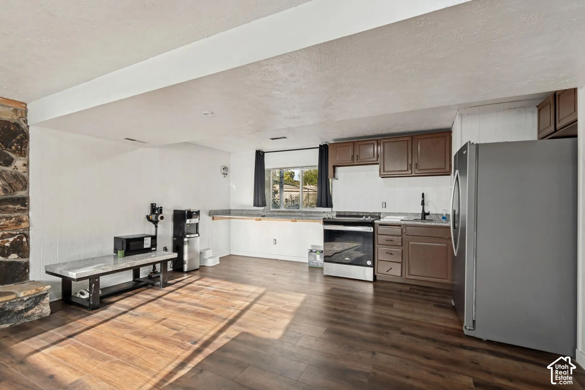Wood-finish flooring, stone-clad column, kitchen area with brown cabinetry, stainless steel range, and stainless steel refrigerator