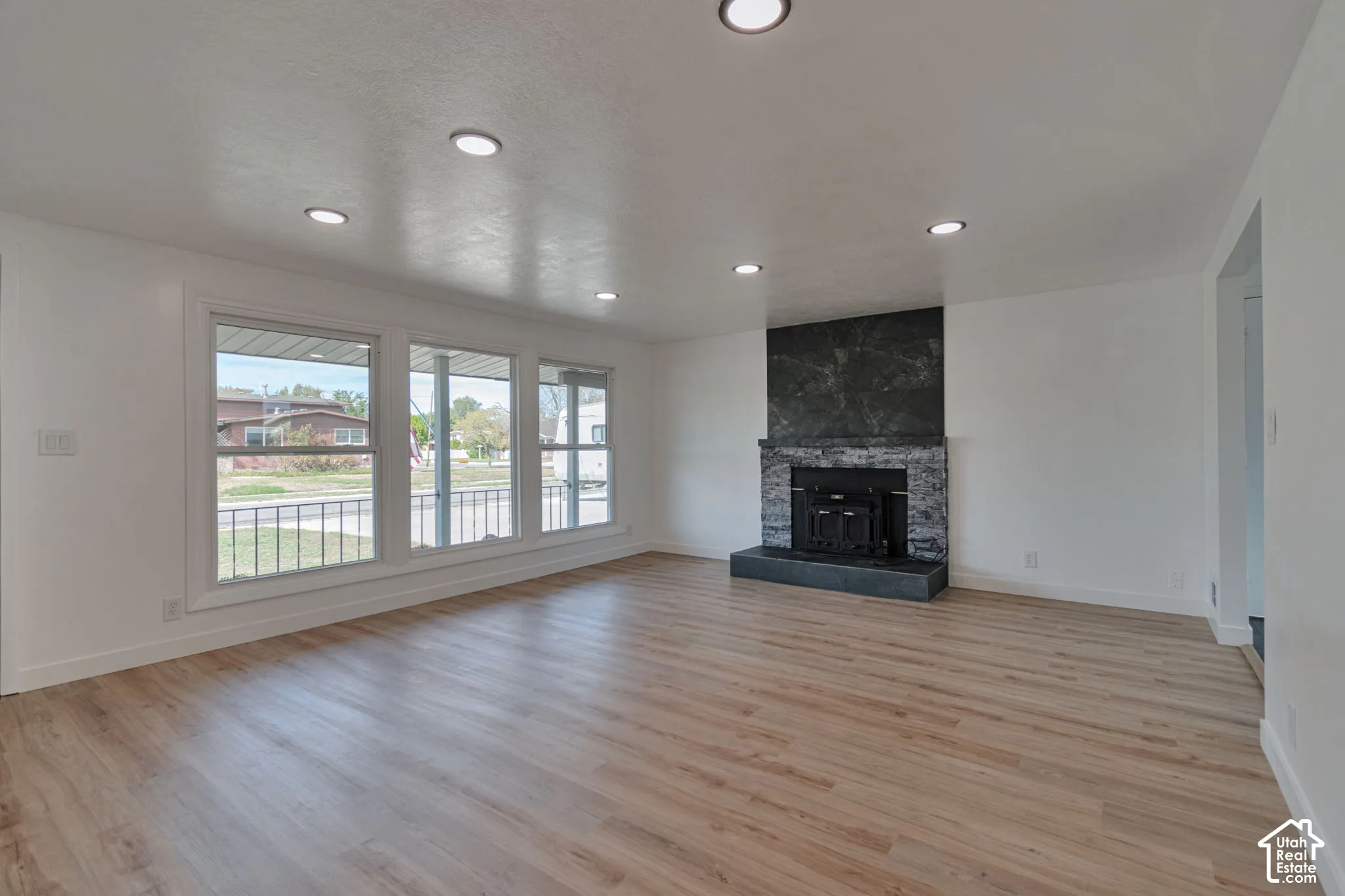 Wood-finish flooring, recessed lighting, white painted walls, a stone-clad fireplace with a dark hearth, and a large window array