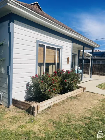 White horizontal siding with blue trim accents, a single-pane window with interior shades, a wooden raised garden bed with shrubbery, and a concrete pathway