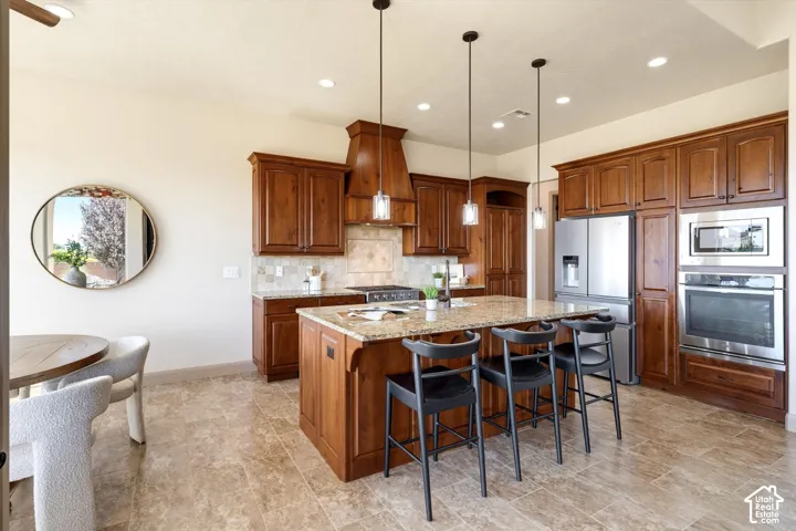 Kitchen featuring wood cabinetry, stainless steel appliances, a multi-toned tile backsplash, and a large central island with a stone countertop