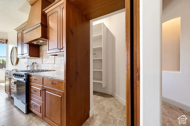 Galley kitchen showcasing rich wood cabinetry with brushed metal hardware, stainless steel gas range and hood, subway tile backsplash, light stone countertops, and wood-finish flooring