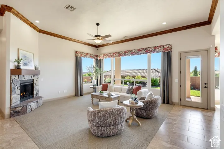 Spacious living area featuring a corner stone fireplace with a wood-finish mantel, large picture windows, a ceiling fan, and crown molding