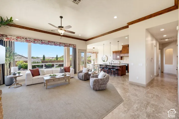 Open-concept living area featuring expansive windows framing scenic vistas, neutral stone flooring throughout, coffered ceiling with wood trim detailing, ceiling fan with integrated lighting, and seamless sightlines to a chef's kitchen with dark cabinetry