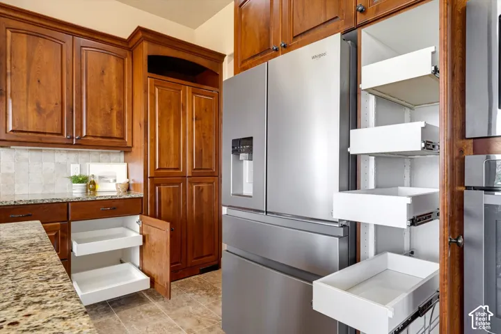Wood-finish cabinetry with crown molding, stainless steel refrigerator, stone-finish countertops, tile backsplash, and pull-out pantry shelving