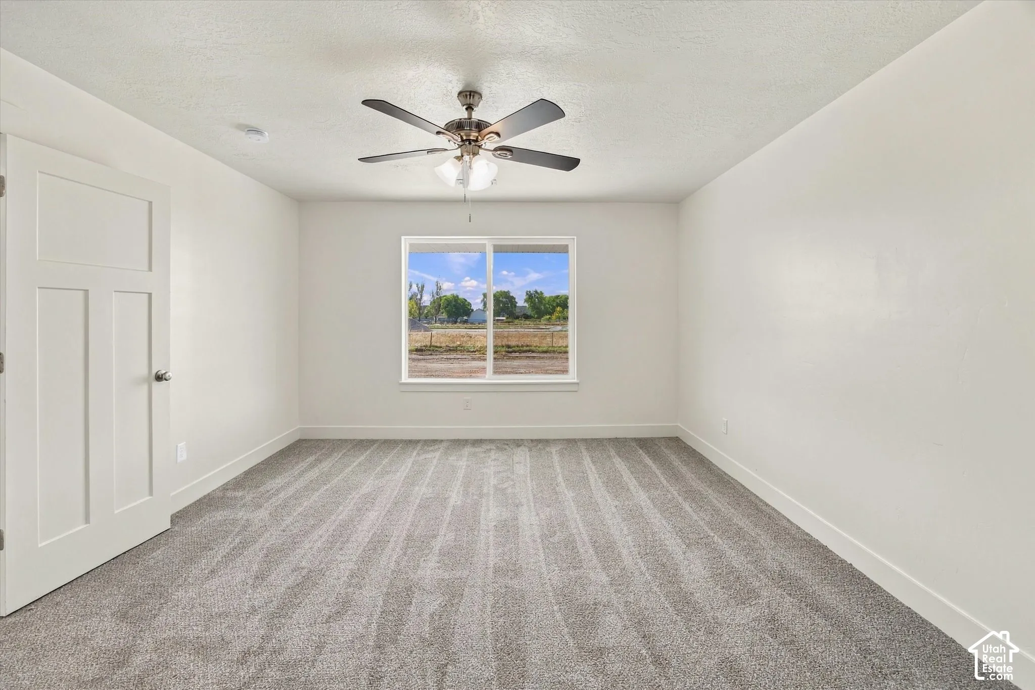 Spacious room featuring gray patterned carpeting, a centered ceiling fan, and a large window with white trim