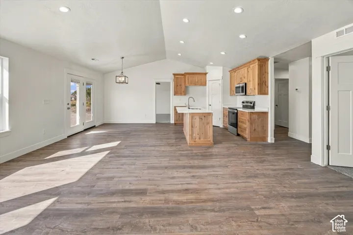 Open-concept living area featuring vaulted ceilings, wood-finish flooring, a kitchen island with a sink, stainless steel appliances, and French doors