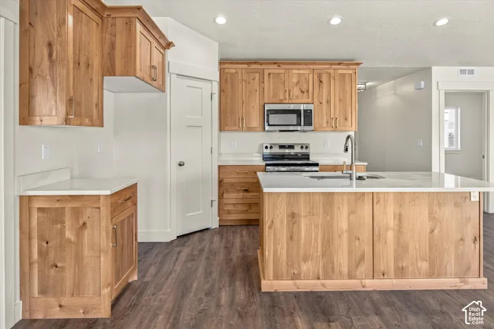 Kitchen featuring wood cabinetry, white countertops, stainless steel appliances, a center island with an integrated sink, and wood-finish flooring
