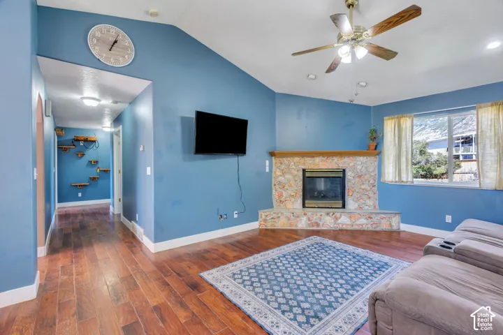 Vaulted ceiling with a ceiling fan, stone-surround fireplace, wood-finish flooring, and a large window