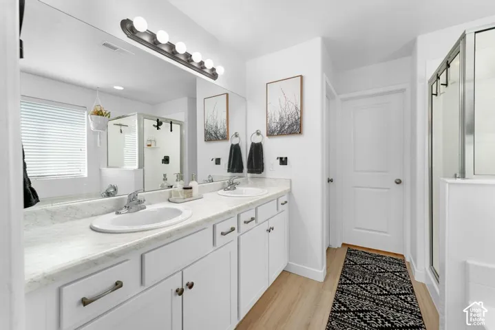 Dual vanity with integrated sinks, full-width mirror, light wood-finish flooring, and a glass-enclosed shower