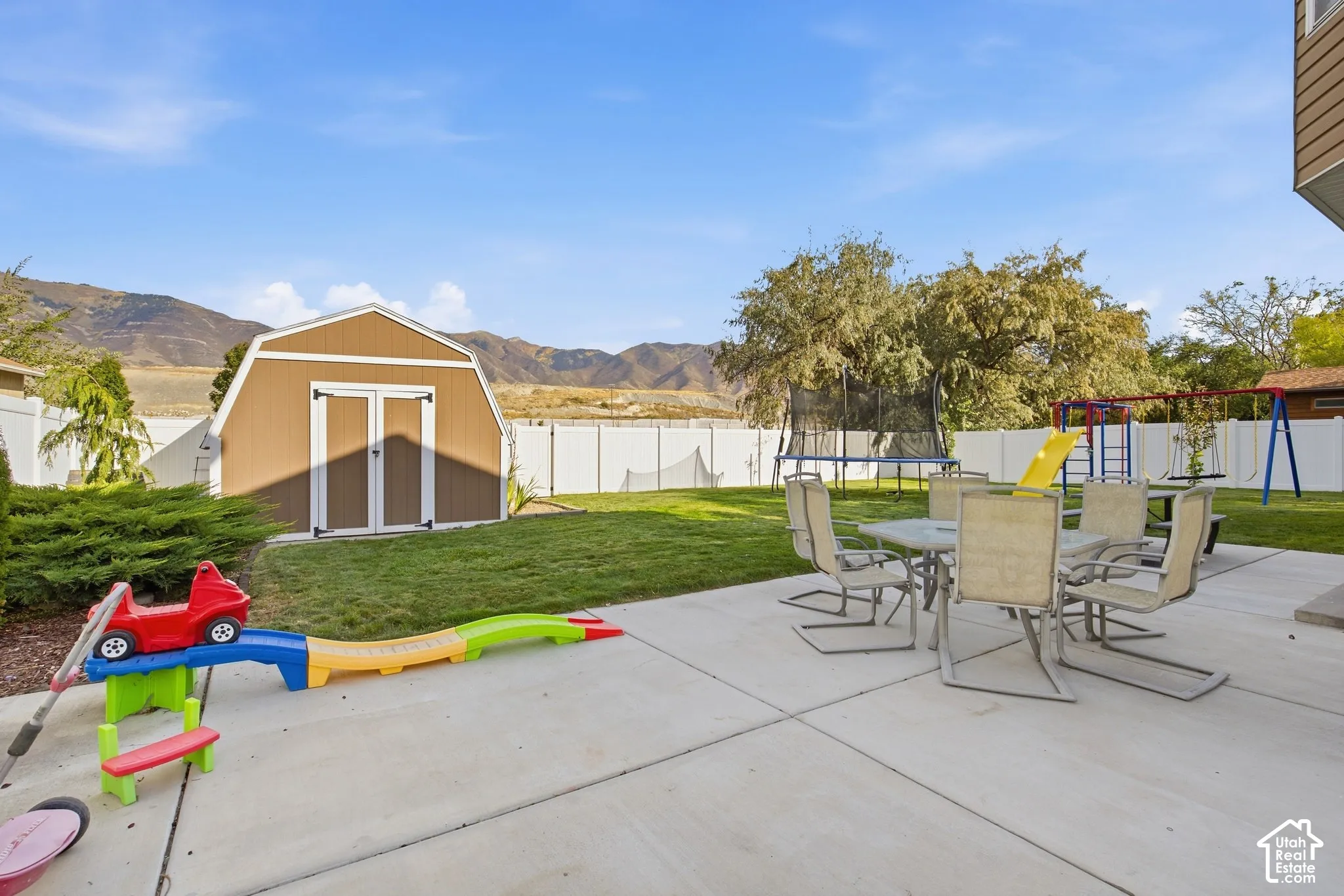 Expansive concrete patio with mature shade trees and manicured lawn, featuring a detached storage shed with peaked roof and white trim, children's play equipment, and patio seating area with mountain views in the distance