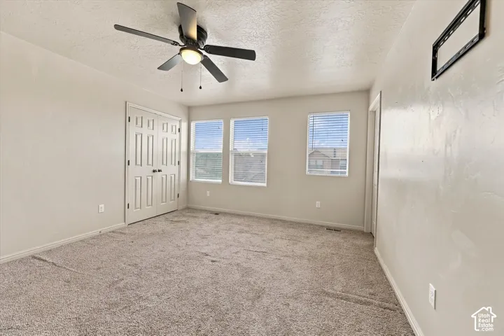 Neutral tone walls, plush carpeting, ceiling fan with integrated lighting, multiple windows with blinds, and paneled bifold closet doors