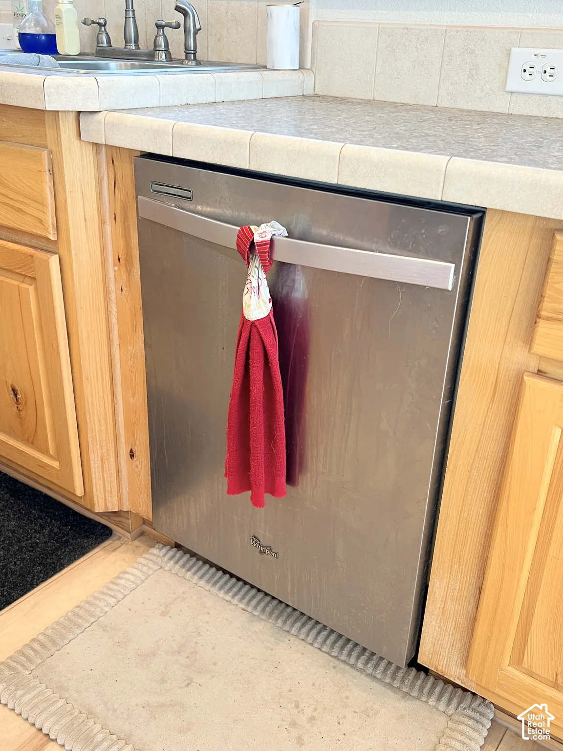 Stainless steel dishwasher, natural wood cabinetry, tiled countertop, and an integrated sink with a dual-handle faucet